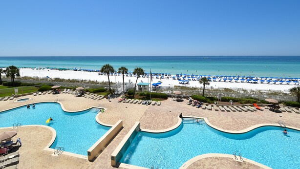 Ein Poolbereich mit mehreren Schwimmbecken, ein Strand mit Sonnenschirmen und einem strahlend blauen Himmel.