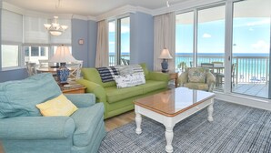 Ein geräumiges Wohnzimmer mit einem grünen Sofa, einem blauen Sofa und einem Couchtisch aus Holz. Ein großes Fenster bietet Blick auf den Ozean.