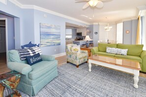 A living room with a teal armchair, a green sofa, a coffee table, and a floral armchair. There is a painting on the wall and a kitchen in the background.