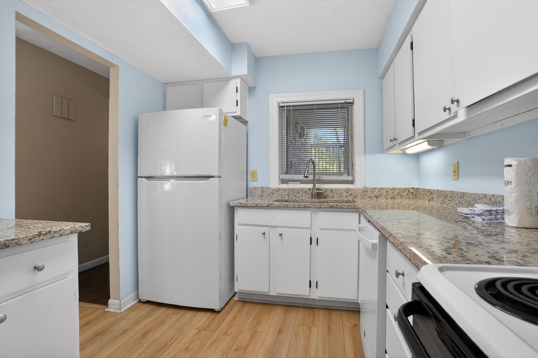 A compact kitchen with white cabinets, a built-in oven, a refrigerator, and a window with blinds.