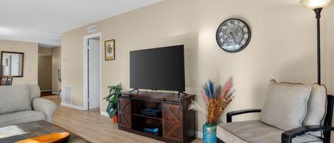 Una sala de estar con una televisión de pantalla plana, una mesa de centro de madera, un sofá, un sillón y un jarrón decorativo.