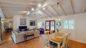 A spacious living area with a high ceiling, a fireplace, a dining table with chairs, and a large window with blinds.