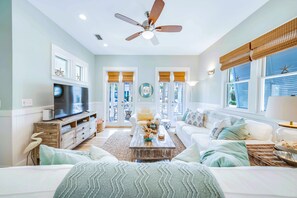 A living room with a television, a ceiling fan, and a large window with blinds.