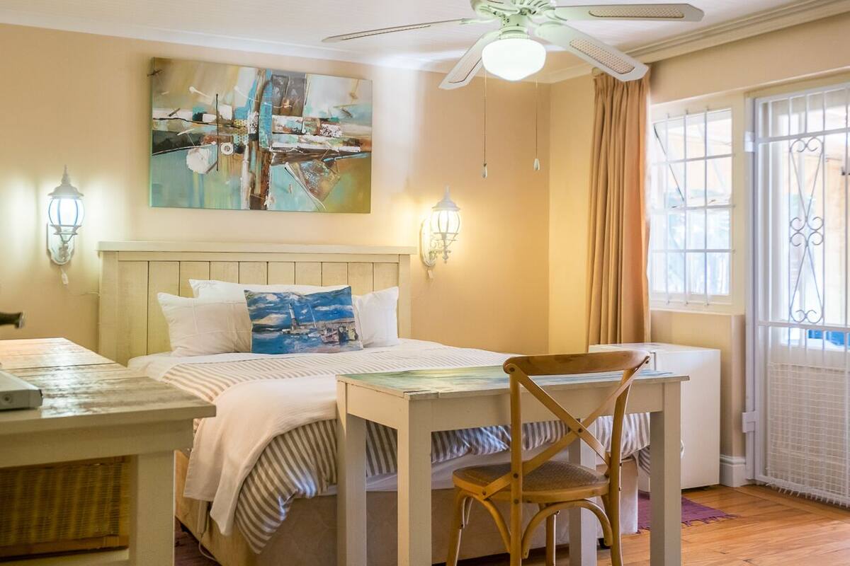 A bedroom with a bed, a table, and a chair. There is a painting on the wall, a ceiling fan, and a window with curtains.