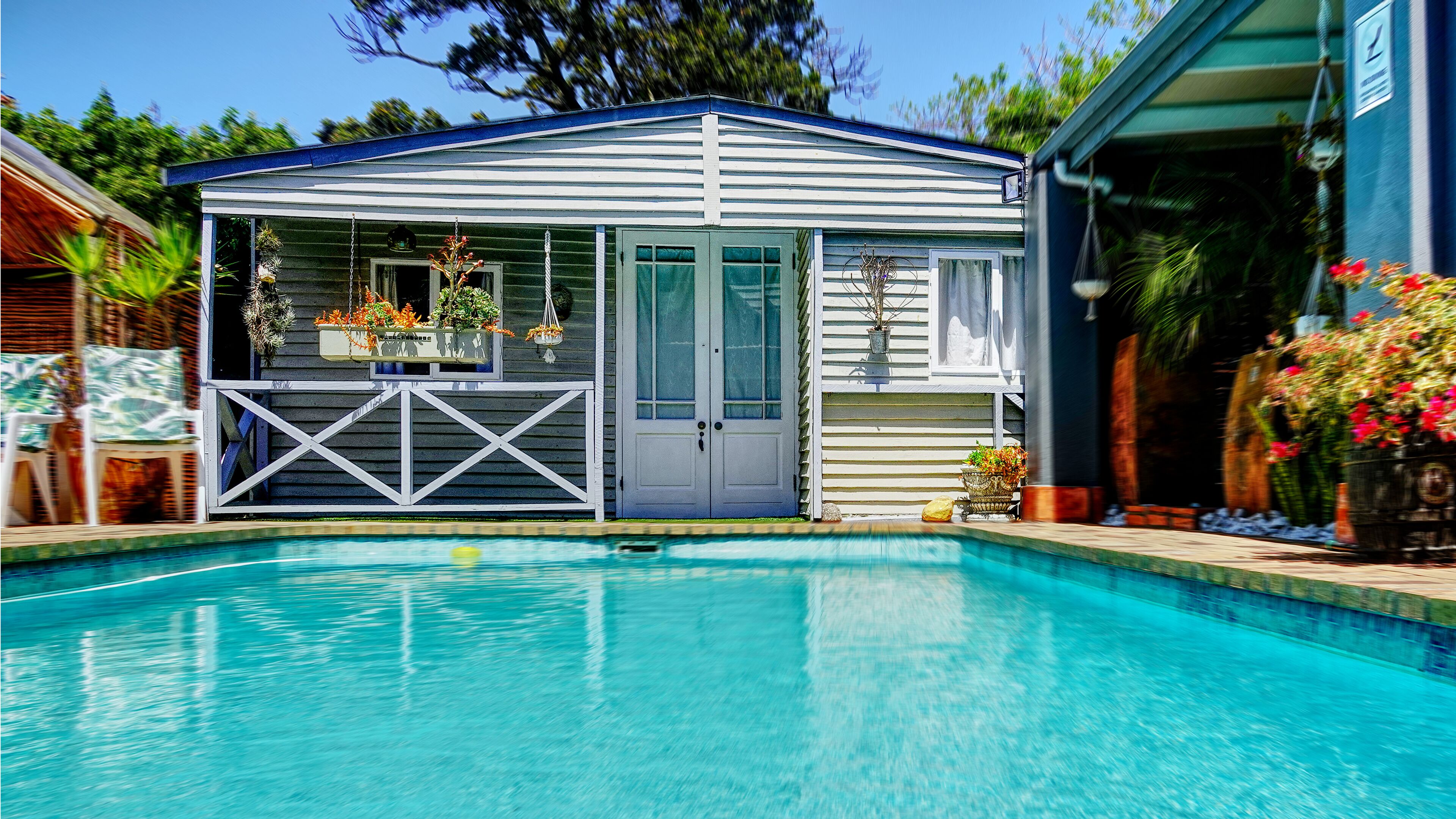 A pool with a covered area featuring a door and windows, surrounded by plants and flowers.