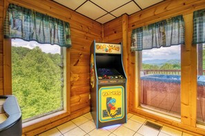 Une borne d’arcade Pac-Man dans une pièce aux murs en bois et aux grandes fenêtres donnant sur une zone boisée.