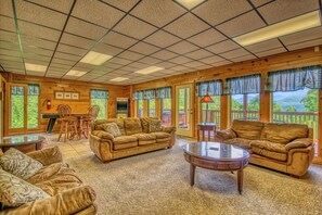 Ein großzügiges Wohnzimmer mit Holzboden, einer großen Couch, einem Couchtisch und einem Kamin. Große Fenster mit Vorhängen bieten einen Blick ins Freie.