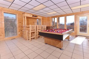 A wooden room with a pool table, chairs, and large windows.