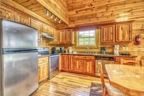 A wooden cabin kitchen with a stainless steel refrigerator, wooden cabinets, a wooden dining table, and a window with a view of greenery.
