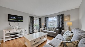 Una sala moderna con un televisor de pantalla plana, un mueble blanco para entretenimiento, un sofá gris y un cojín decorativo estampado.