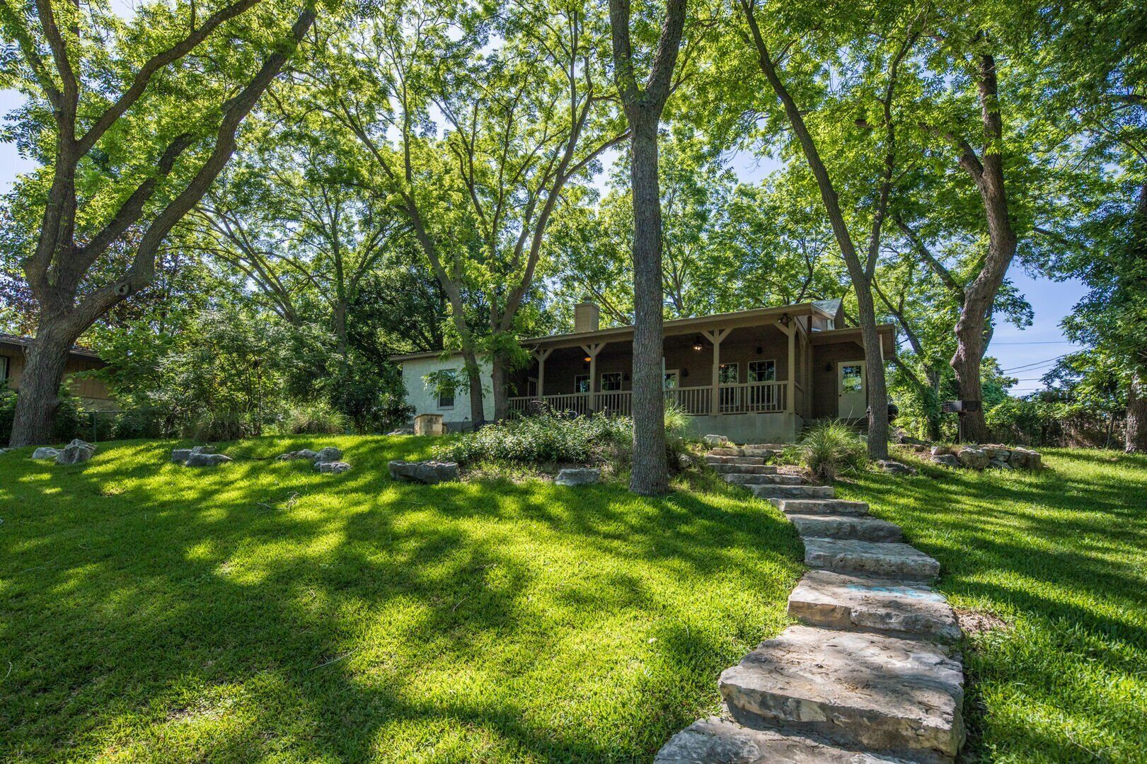 A house with a stone pathway leading to a porch, surrounded by lush greenery and trees.