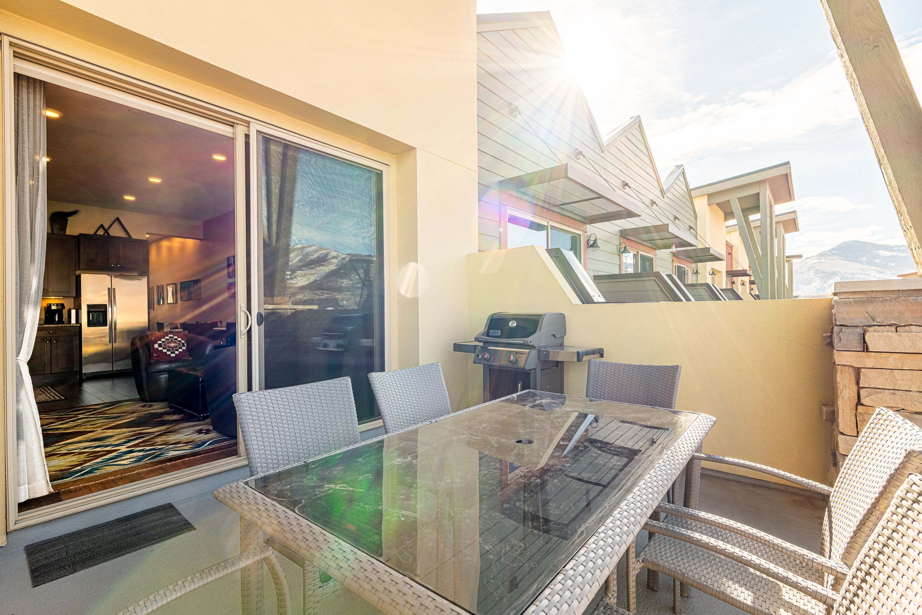 A glass-top dining table with wicker chairs, a grill, and a view of a mountain.