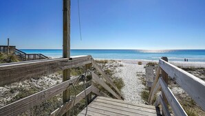 Ein hölzerner Steg führt zu einem Strand mit blauem Himmel und dem Meer im Hintergrund.
