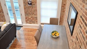 Un coin repas avec une table en bois, des chaises et un saladier rempli de fruits. Il y a un mur en briques et une fenêtre avec des stores.
