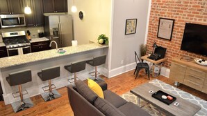 Une cuisine moderne dotée d’appareils électroménagers en acier inoxydable, un coin bar avec des tabourets, un espace salon avec un canapé et une table basse, et un mur en briques avec un téléviseur.