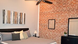 Une chambre avec un mur en briques, un lit avec une literie grise, un ventilateur de plafond et une commode avec un miroir.