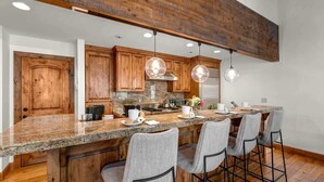 A kitchen with wooden cabinets, a granite countertop, bar stools, pendant lights, and a stainless steel range hood.