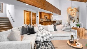 A modern living room with a sectional sofa, wooden beams, a staircase, and a kitchen in the background.