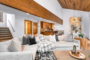 Uma sala de estar moderna com sofá seccional, vigas de madeira, uma escada e uma cozinha ao fundo.
