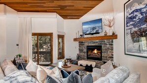 A cozy living room with a stone fireplace, a television mounted above it, and a large window overlooking a deck.