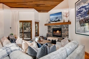 Uma sala de estar aconchegante com lareira de pedra, uma televisão montada acima dela e uma janela grande com vista para um deck.