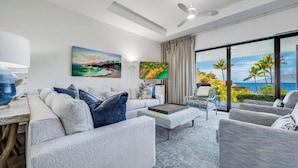 A modern living room with a sofa, armchairs, and a coffee table. There is a painting on the wall, a TV, and a view of the ocean through large windows.