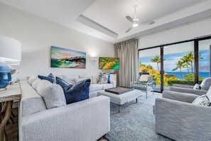 Un salon moderne avec un canapé, des fauteuils et une table basse. Un tableau orne le mur, il y a un téléviseur et, grâce à de grandes fenêtres, on peut admirer la vue sur l’océan.