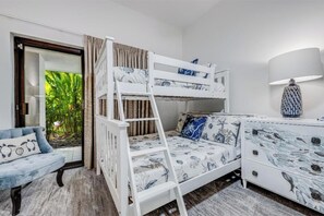 A bunk bed with a ladder, a blue sofa, a white dresser with a marine design, and a view of greenery through a glass door.