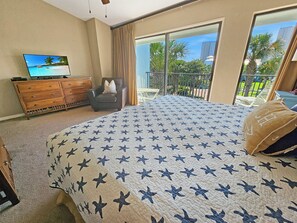 Une chambre avec un grand lit, un téléviseur posé sur une commode en bois, et un balcon offrant une vue sur les palmiers et les immeubles.