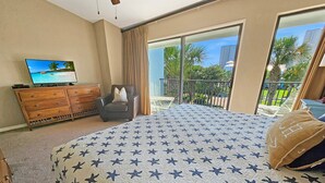 Quarto com cama grande, TV em cômoda de madeira e varanda com vista para palmeiras e edifícios.