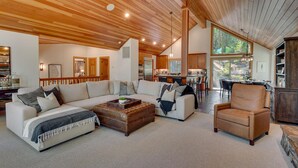 A spacious living room with a large sectional sofa, a leather ottoman, and a leather armchair. A kitchen is visible in the background with wooden cabinets and a dining area. - Tahoe Treehouse - TLUXP (Homewood)