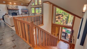 A spacious living area with a stone fireplace, wooden staircase, and large windows offering a view of the outdoors. - Tahoe Treehouse - TLUXP (Homewood)