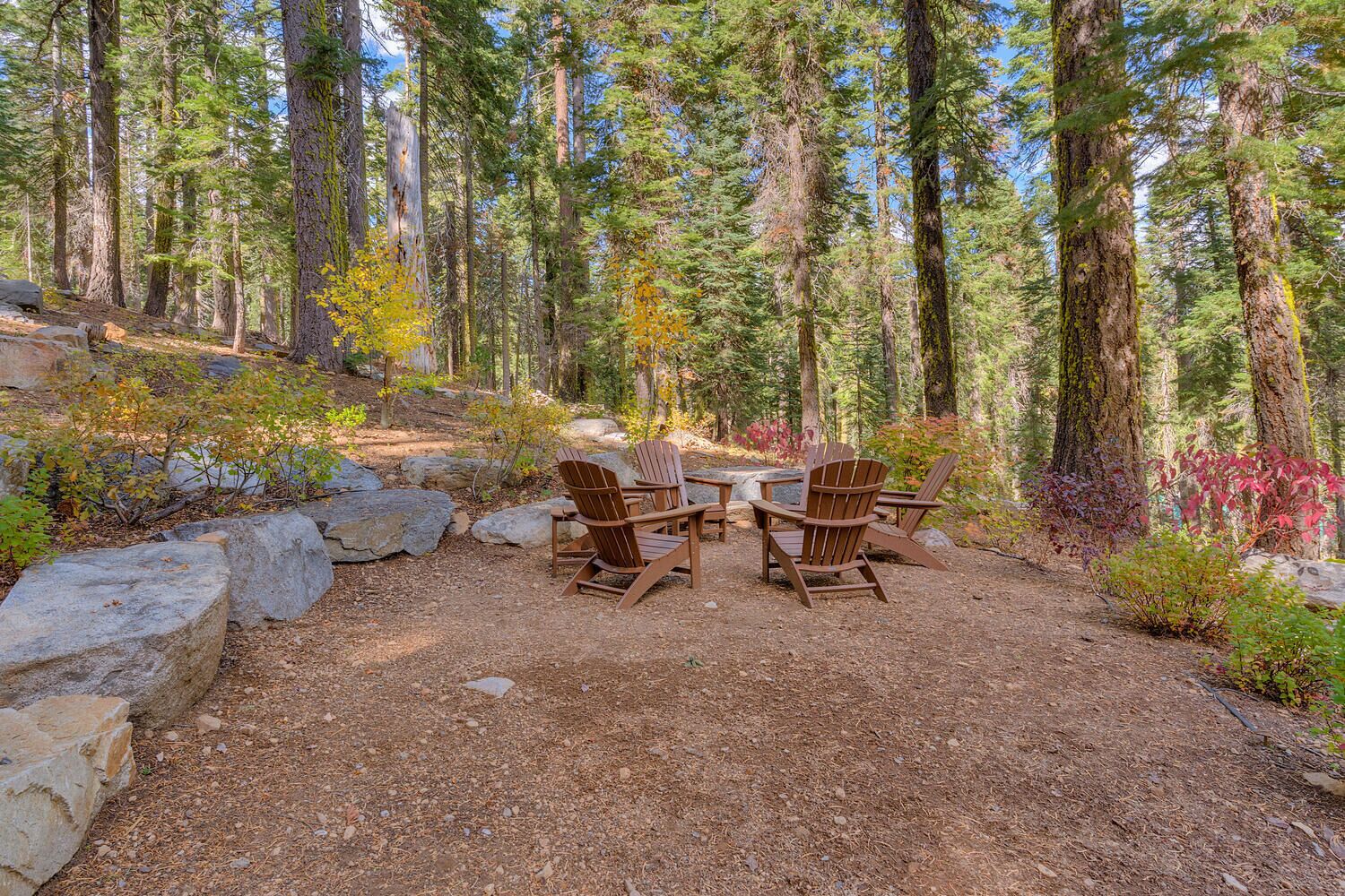 A forested area with a seating arrangement of wooden chairs and tables, surrounded by rocks and trees.
