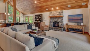 A spacious living room with a stone fireplace, a large sofa, and a television. - Tahoe Treehouse - TLUXP (Homewood)
