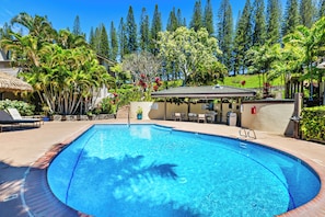 A swimming pool with a sunshade, lounge chairs, and a covered seating area surrounded by tropical plants and trees.