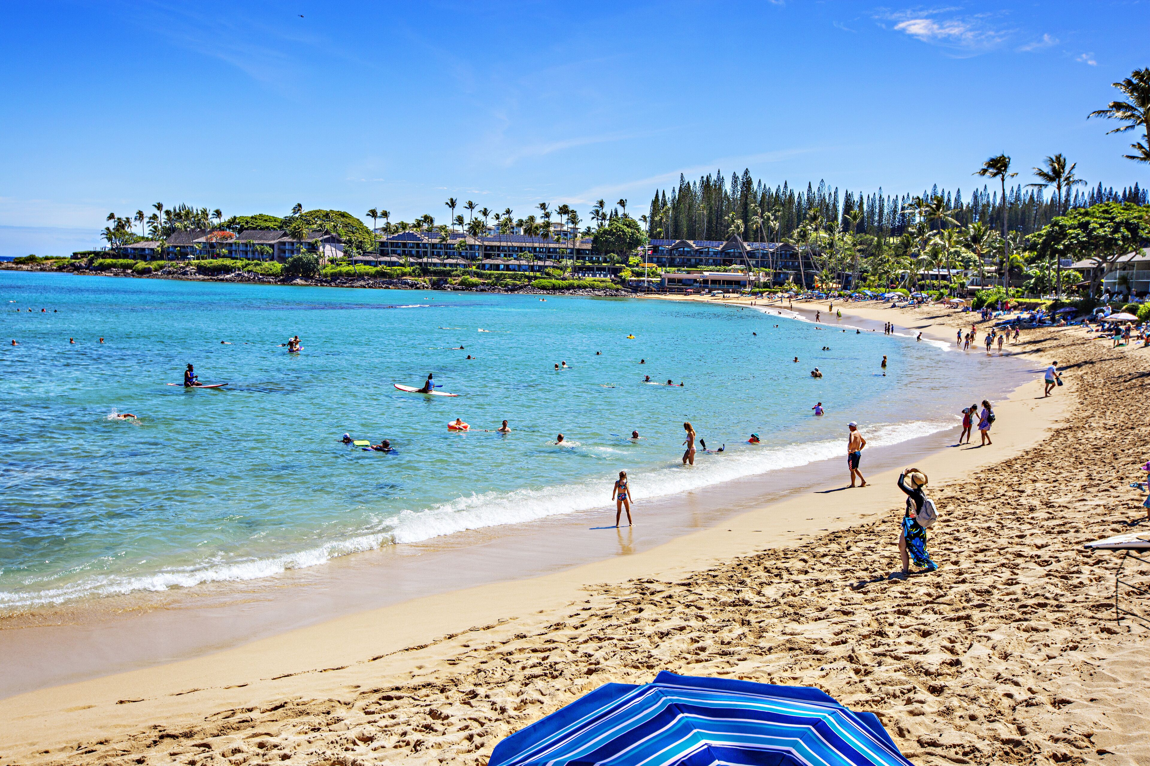Ein Strand mit klarem türkisblauem Wasser, Menschen beim Schwimmen und Spazierengehen im Sand, und ein Resort im Hintergrund.