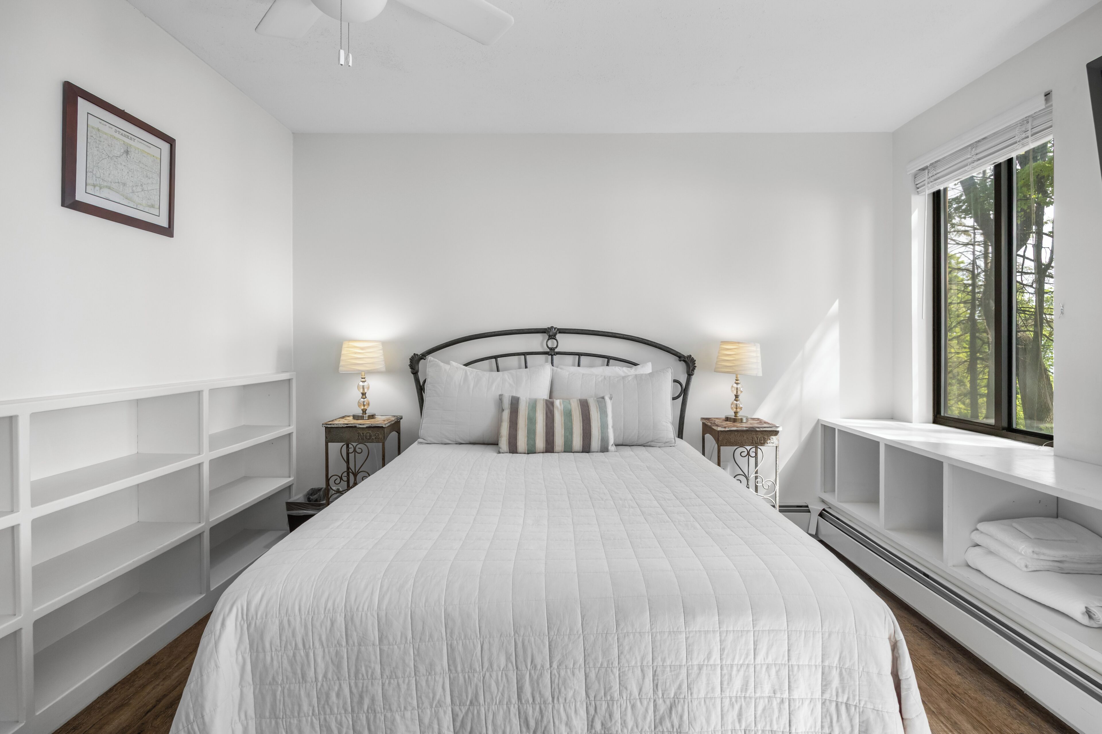 A bedroom with a bed, two bedside tables with lamps, a window with a view, and built-in shelving.