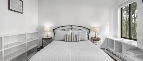 A bedroom with a bed, two bedside tables with lamps, a window with a view, and built-in shelving.