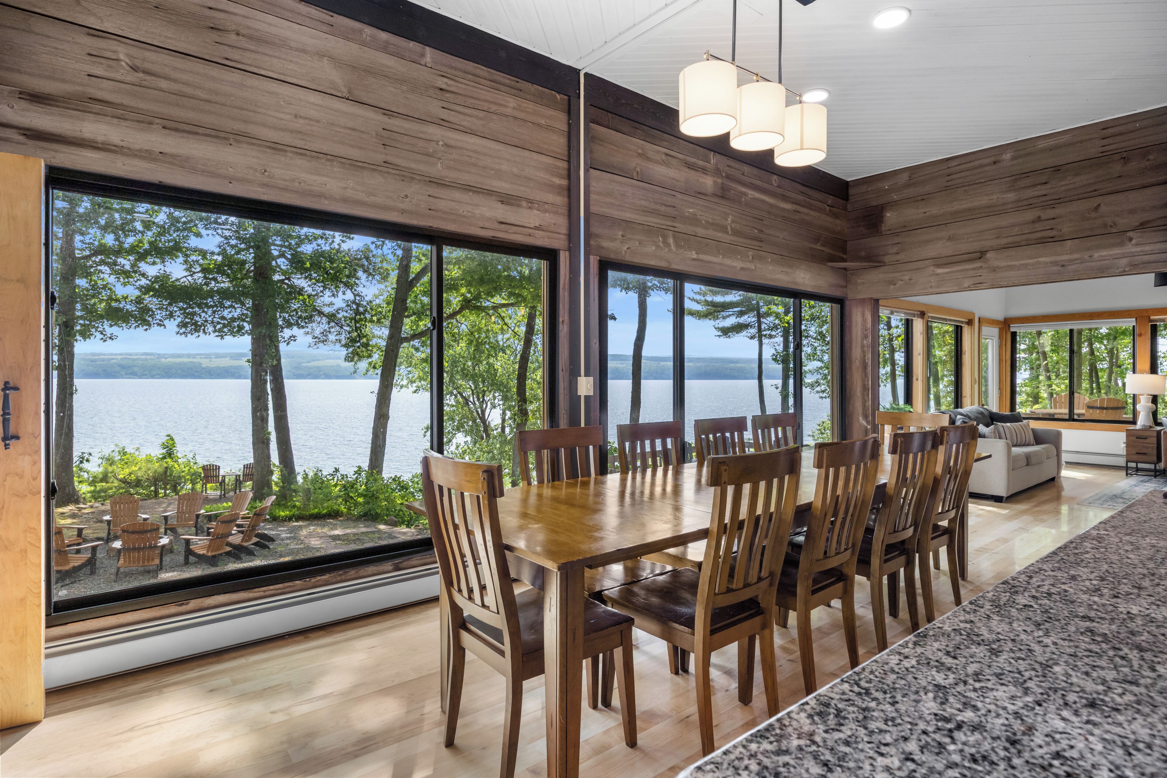 A dining area with a large table and chairs, large windows with a view of trees and water, and a modern kitchen area.