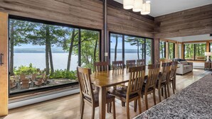 Un espace repas comprenant une grande table et des chaises, de grandes fenêtres offrant une vue sur les arbres et l’eau, ainsi qu’une cuisine moderne.