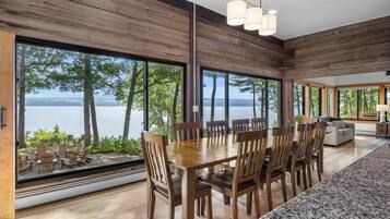 A dining area with a large table and chairs, large windows with a view of trees and water, and a modern kitchen area.
