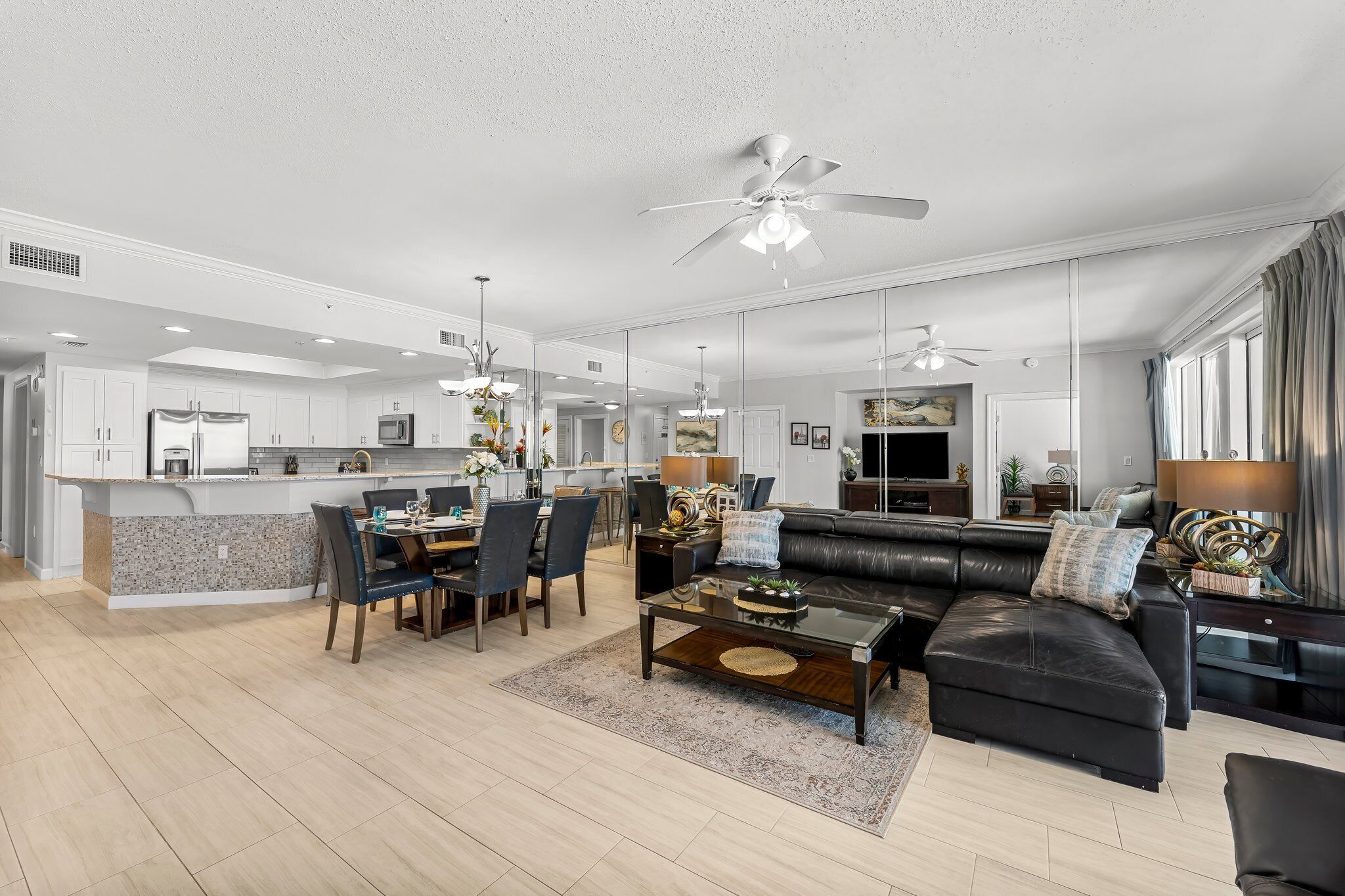 A spacious living area with a black leather sofa, a dining table with chairs, and a kitchen area with white cabinets and a white ceiling fan.