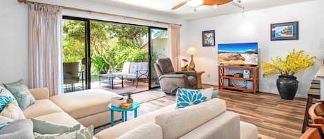 A living room with a sofa, armchairs, a coffee table, and a TV on a wooden stand. There is a sliding glass door leading to a patio with outdoor furniture.