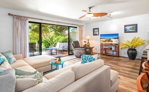 Ein Wohnzimmer mit einer Couch, Sesseln, einem Couchtisch und einem Fernseher auf einem Holzständer. Eine Glastür führt zu einer Terrasse mit Gartenmöbeln.