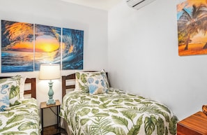A room with two beds, a nightstand with a lamp, and wall art depicting a sunset and palm trees. - Maui Kamaole M-105 Ground Floor, Spacious, Secluded, Full A/C, Great Location (Kihei)