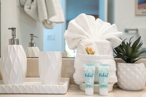 Et badeværelsesbord med et hvidt håndklæde, to flasker Kekaï shampoo og balsam, en potteplante og en hvid sæbedispenser.