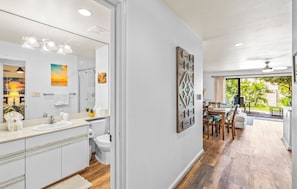 A modern bathroom with a large mirror, sink, and toilet. There is a dining area with a table and chairs visible through an open doorway. - Maui Kamaole M-105 Ground Floor, Spacious, Secluded, Full A/C, Great Location (Kihei)