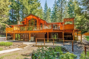 A large wooden cabin with multiple decks and a covered outdoor area, surrounded by dense forest.
