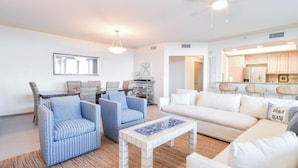 Een ruime woonkamer met een witte bank, twee gestreepe fauteuils, een salontafel en een eethoek met rieten stoelen.