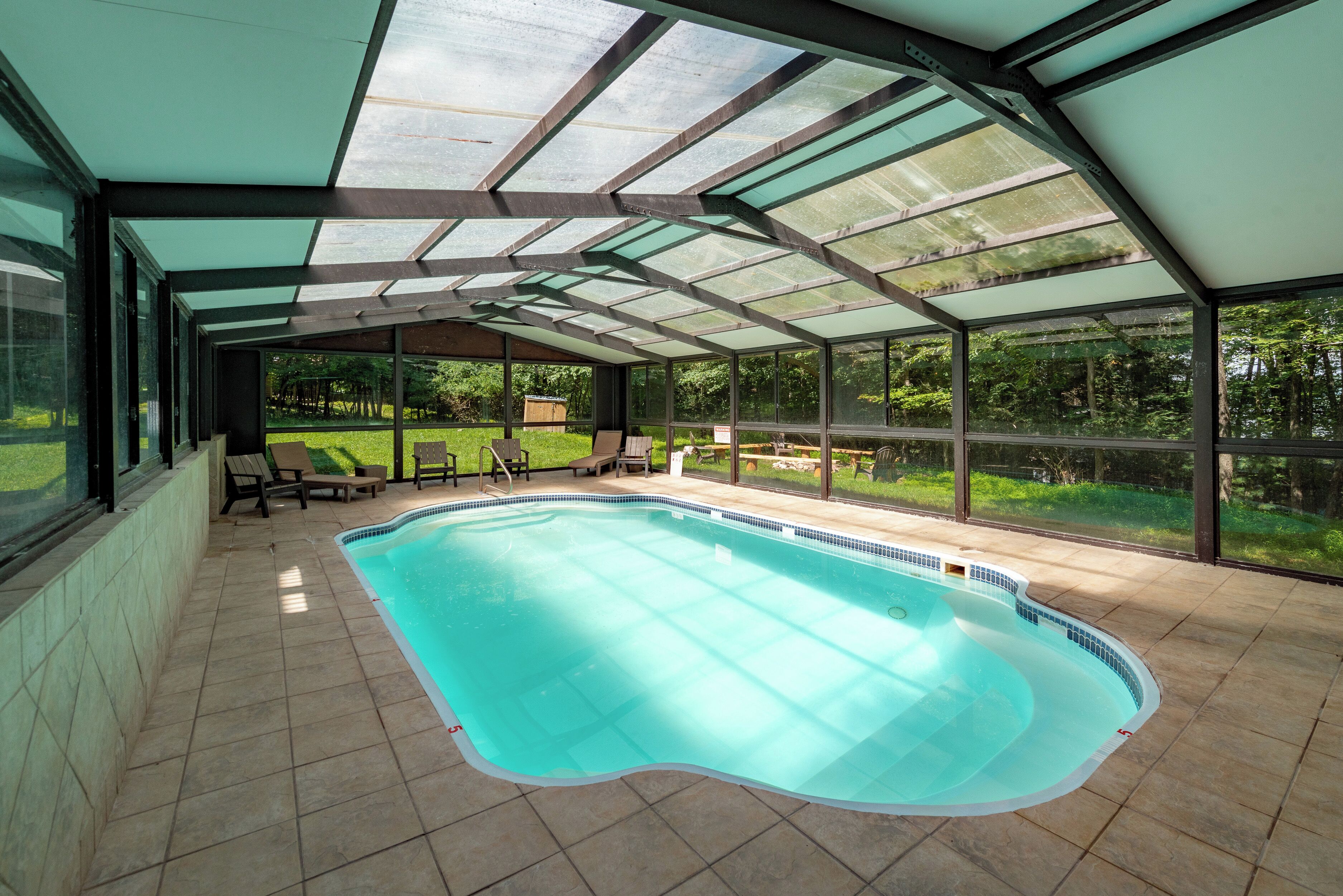 An indoor swimming pool with glass walls and a tiled floor, surrounded by lounge chairs and a view of a green landscape.